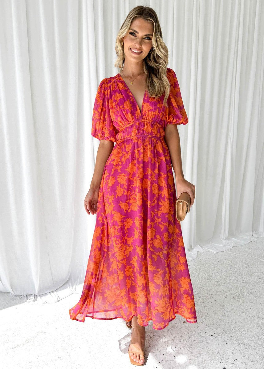 Woman wearing pink and orange floral midi dress with clutch, standing on textured floor with white curtain backdrop.