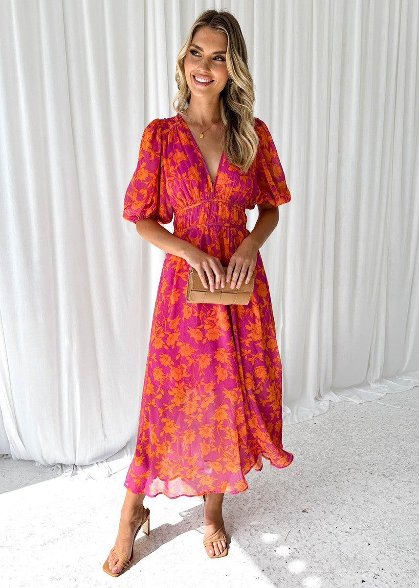 Woman in pink and orange floral midi dress holding clutch, standing on a textured indoor floor with white curtains.