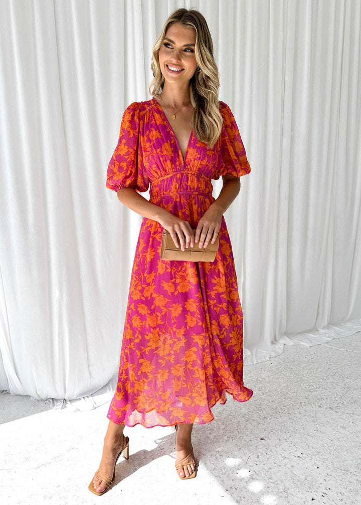 Woman in pink and orange floral midi dress holding clutch, standing on a textured indoor floor with white curtains.