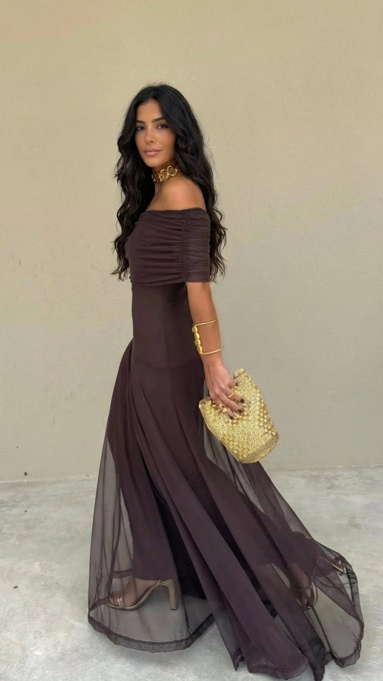 Woman in a flowing off-shoulder brown dress with a sheer overlay, carrying a textured gold clutch, standing against a plain wall indoors.
