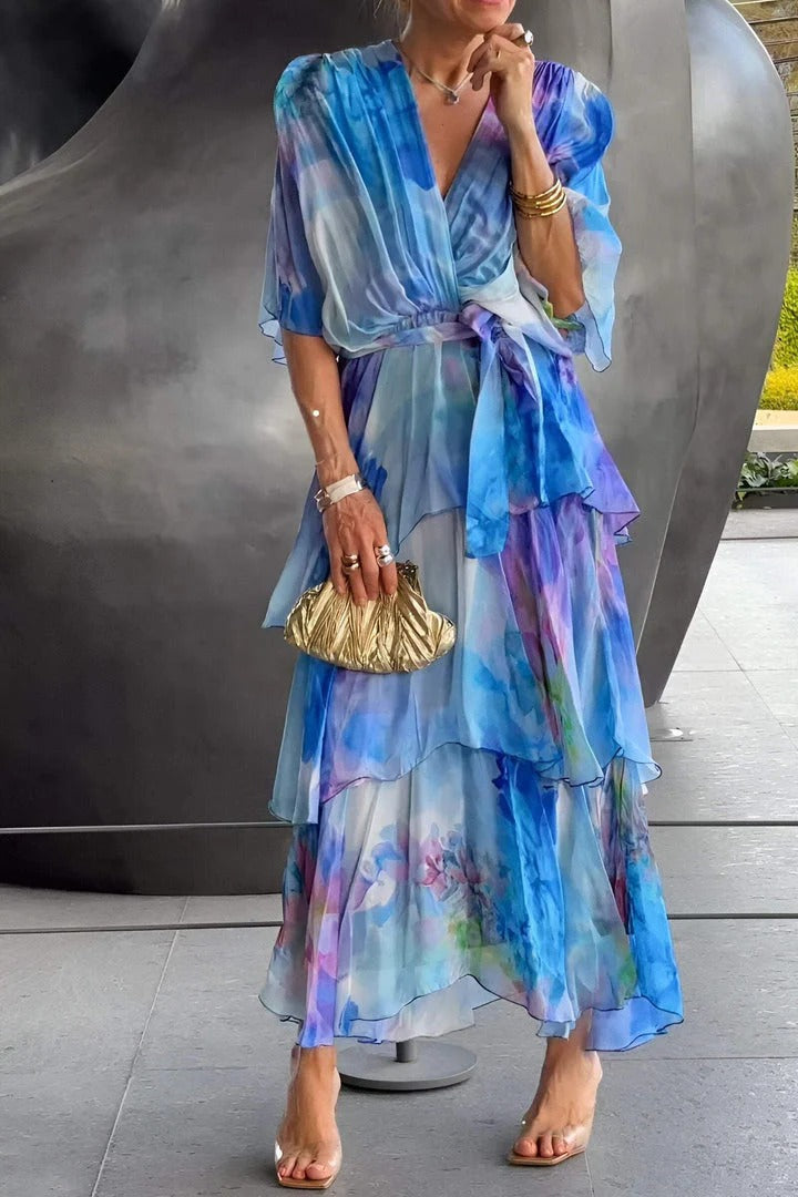 Elegant flowy dress in blue and purple hues, worn by a person holding a gold clutch. Standing indoors against a metallic backdrop.