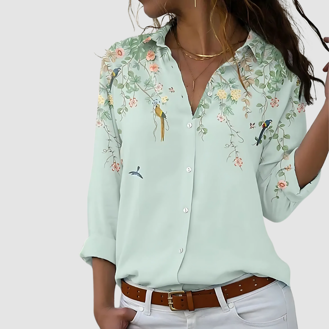 Soft blouse with delicate floral and bird details on a light mint background, paired with white pants indoors.