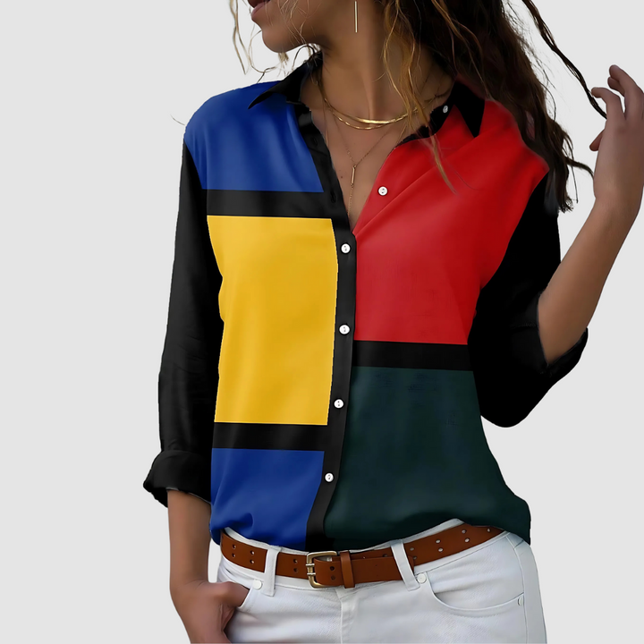 Blouse with bold geometric color blocks in blue, red, yellow, and black, worn with white pants indoors.