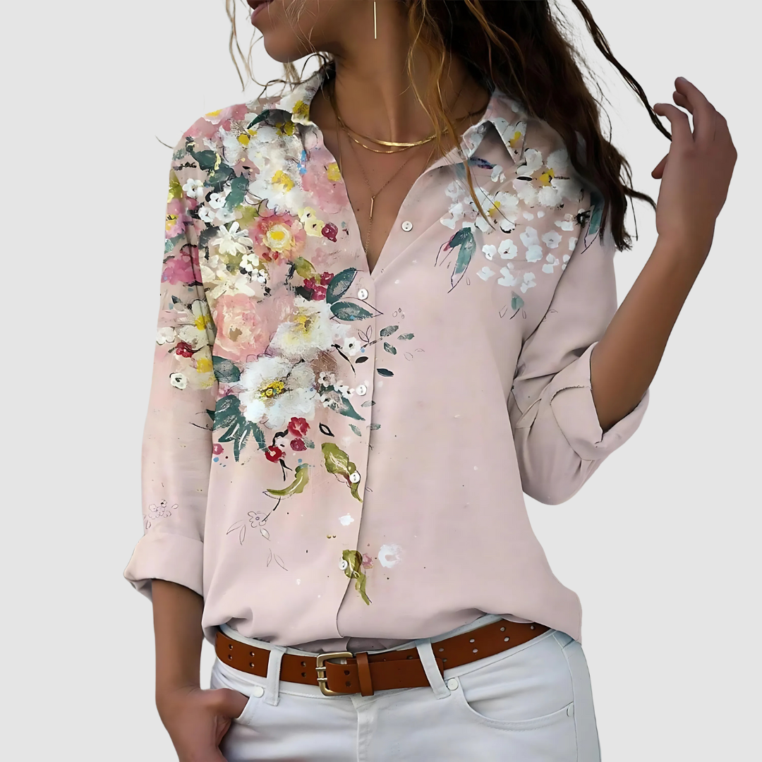 Soft blouse with vivid floral design in pastel pink, worn tucked into white pants against a plain backdrop.