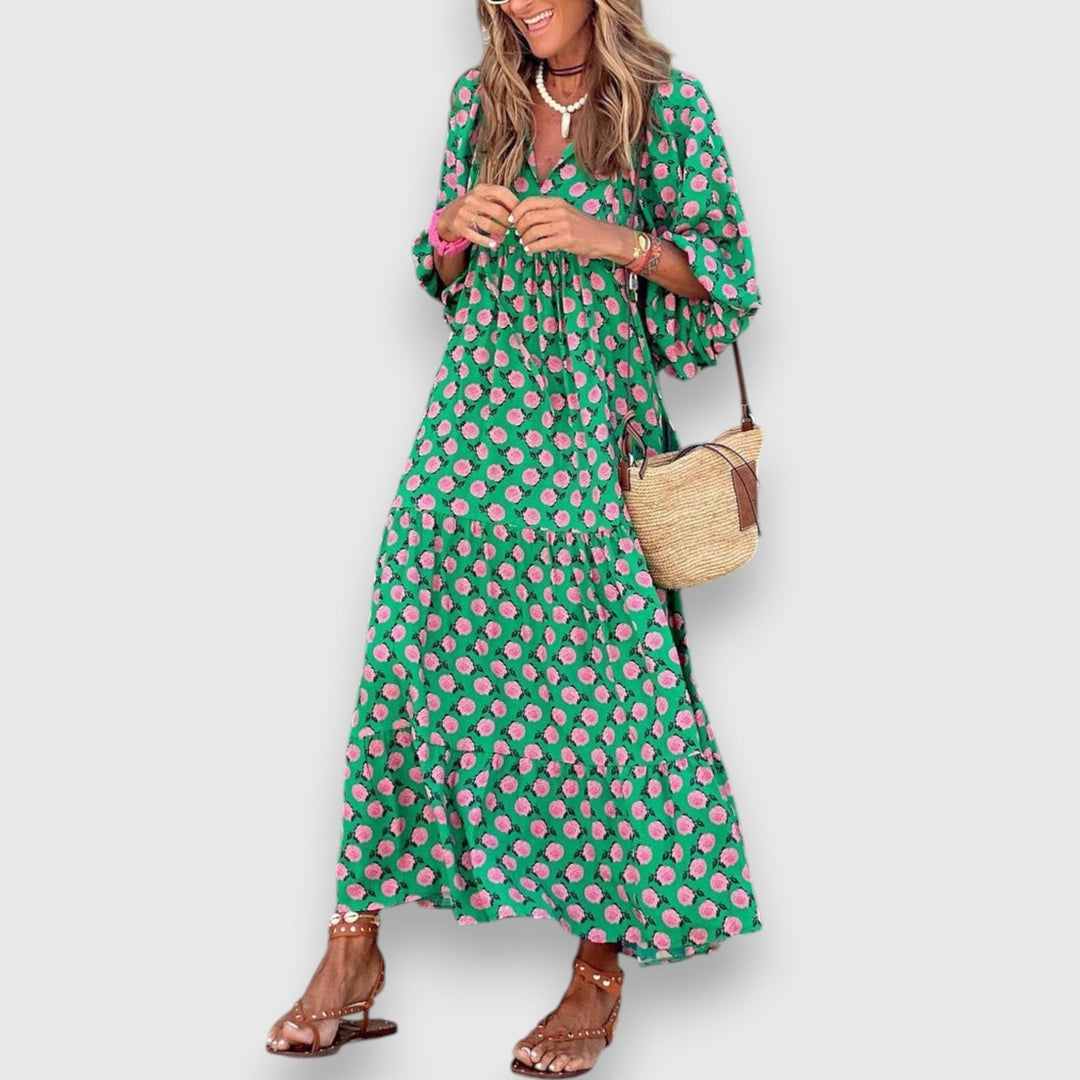 Woman in green dress with pink floral pattern, wearing sandals and holding woven bag, on plain background.