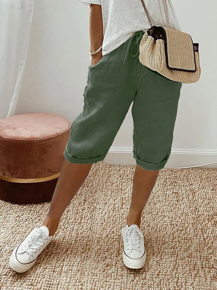 Green comfort shorts styled with a white top and sneakers, displayed in a cozy indoor space with woven rug and stool.