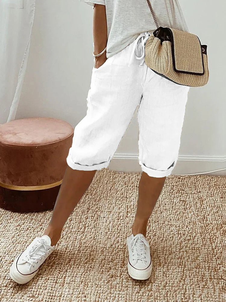 White comfort shorts accompanied by a white top and casual sneakers, set in a relaxed indoor environment with woven rug and stool.