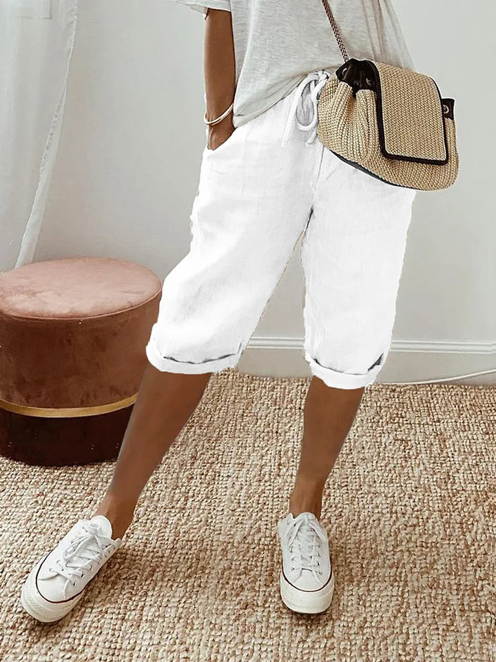 White comfort shorts accompanied by a white top and casual sneakers, set in a relaxed indoor environment with woven rug and stool.