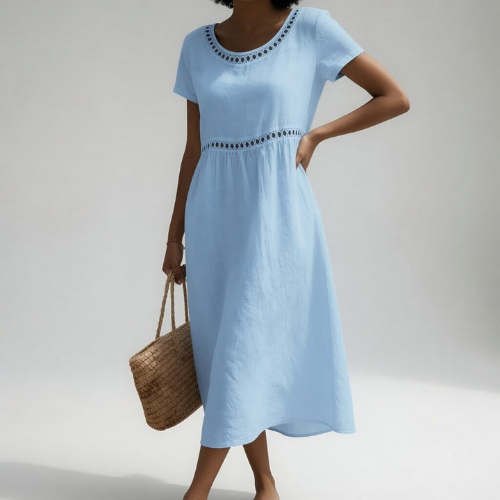 Sky blue midi dress with eyelet detailing and short sleeves, shown on a person holding a woven bag. Clean studio setting.