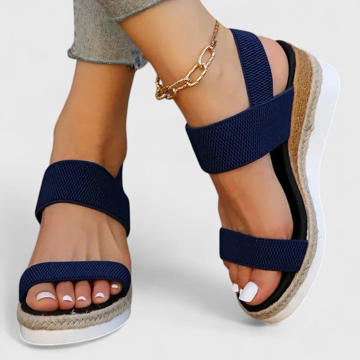 Blue orthopedic sandals with cross straps and wedge soles, worn with light jeans and a gold anklet in a studio setting.