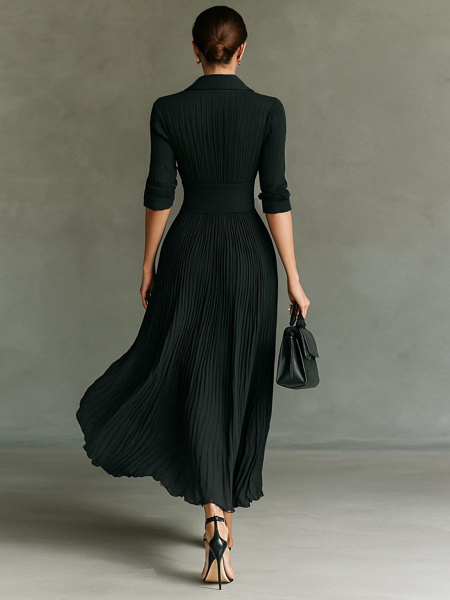 Back view of a black pleated maxi dress worn by a model holding a handbag, walking against a gray studio backdrop.