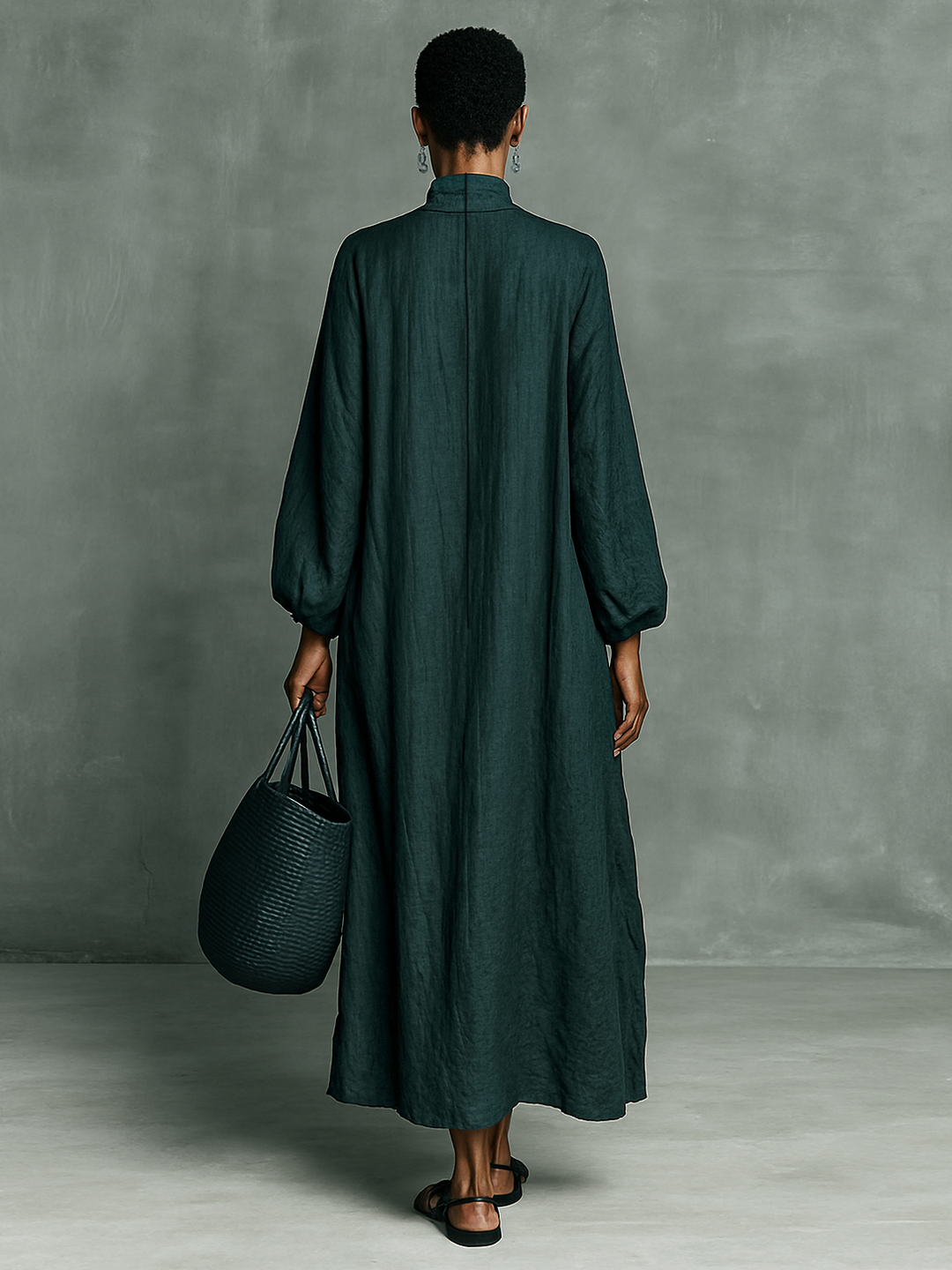Back view of model in green maxi dress holding a woven tote against a minimal studio background.