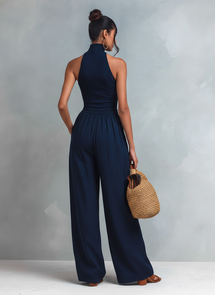 Back view of woman wearing a dark blue jumpsuit, holding a woven bag, in a studio setting.