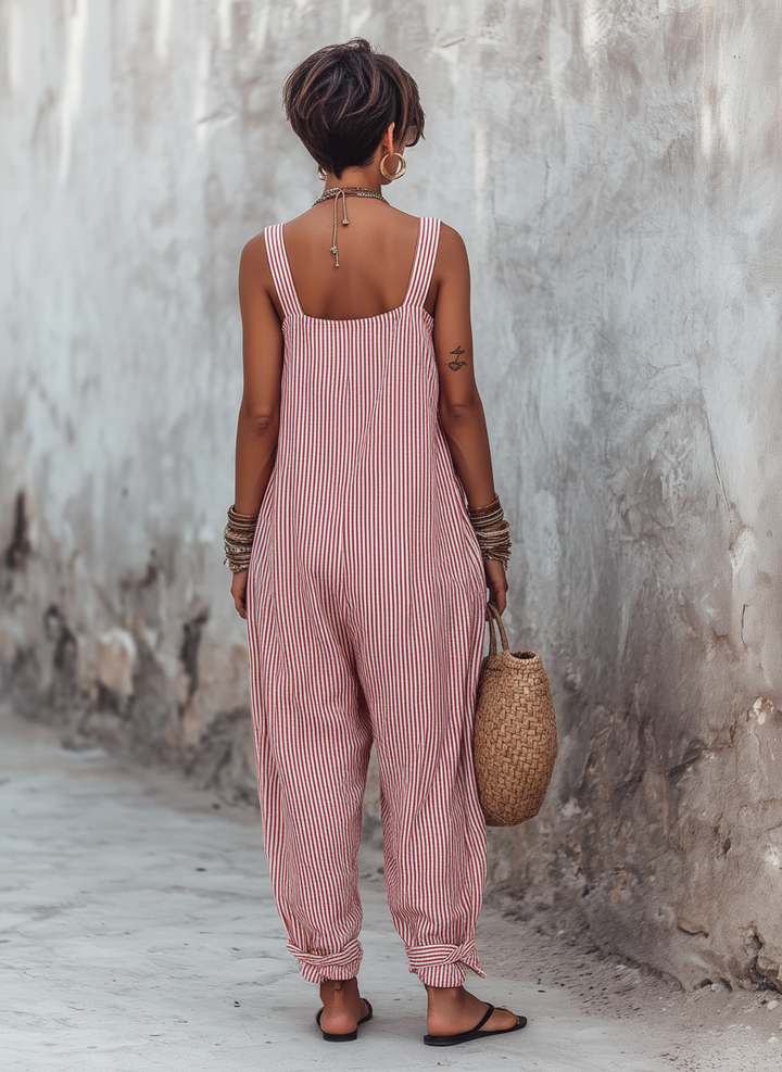 Back view of red and white striped jumpsuit on a woman outdoors, holding a woven bag, with a textured wall background.