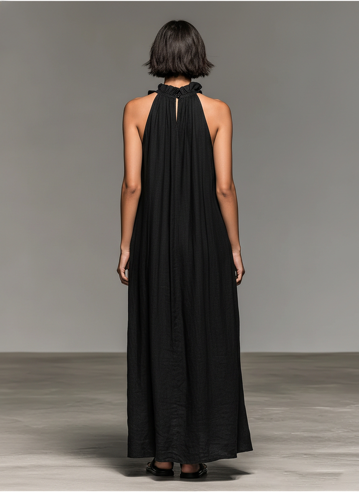 Back view of black maxi dress with ruffled neckline and flowing design, model standing on concrete floor against gray background.