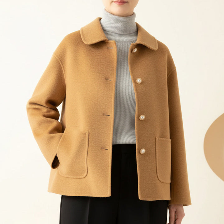 Beige jacket worn over gray turtleneck, hands in pockets, in a neutral room.