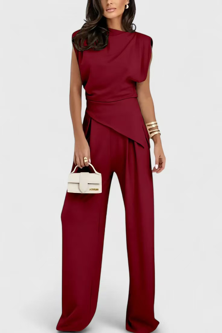 Elegant two-piece burgundy set with draped top and wide-leg pants, styled with gold bangles and white handbag in a studio setting.