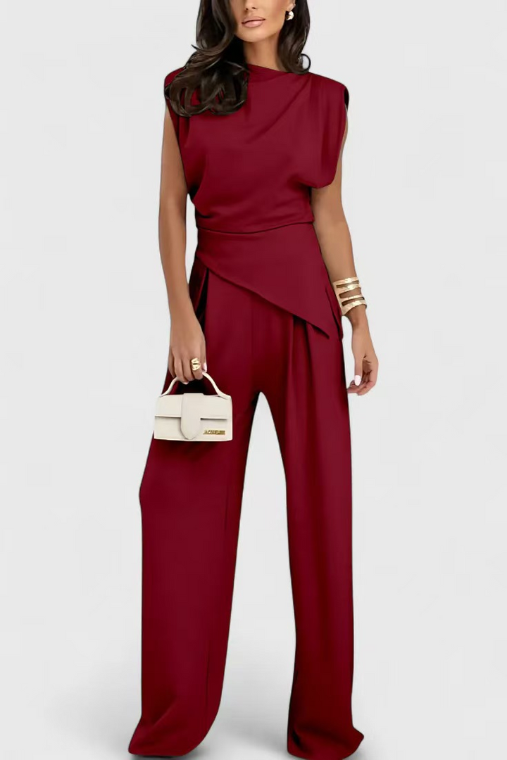Elegant two-piece burgundy set with draped top and wide-leg pants, styled with gold bangles and white handbag in a studio setting.