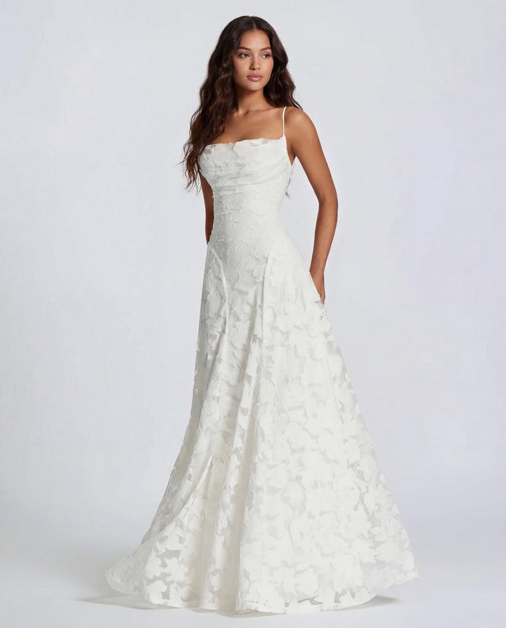 Model in a white maxi dress with thin straps and floral design, studio background.