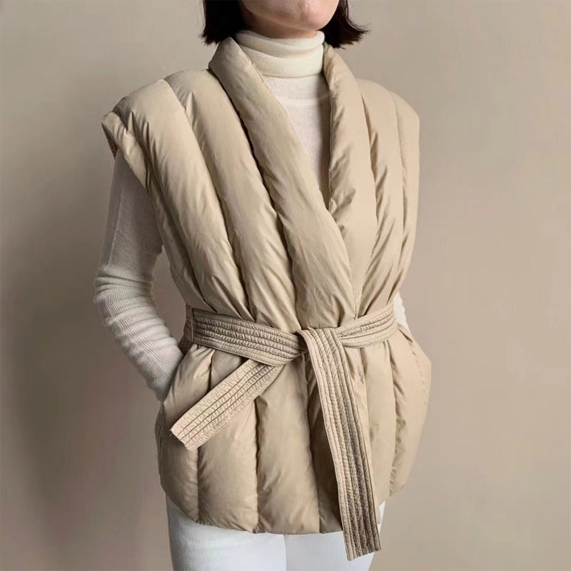 Cream puffer vest with structured wrap and tied belt over a turtleneck. Studio background.