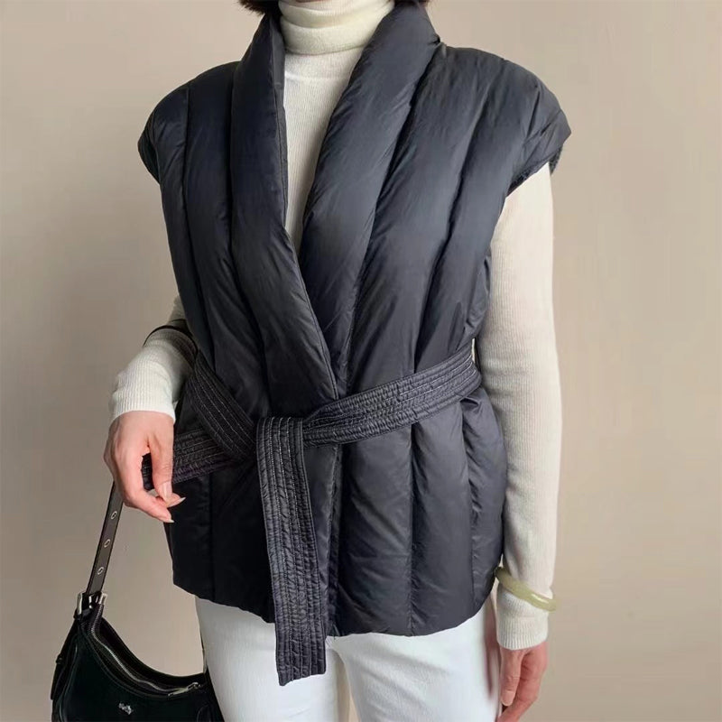 Black puffer vest with belted wrap over a turtleneck, accessorized with a bag. Studio background.