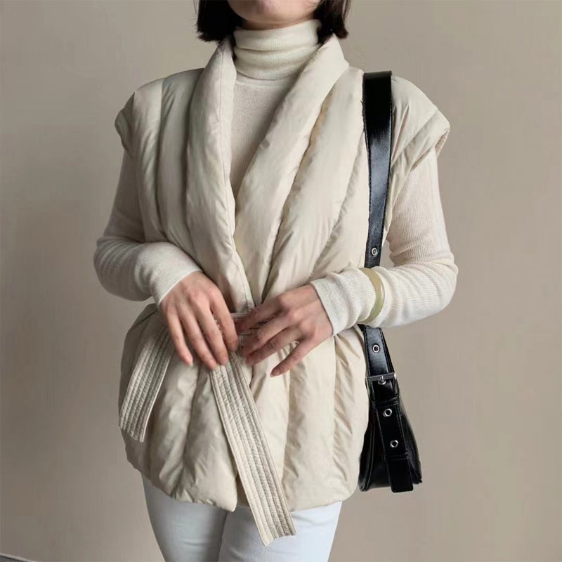 Cream puffer vest with a soft, belted wrap, worn over a turtleneck. Studio background.