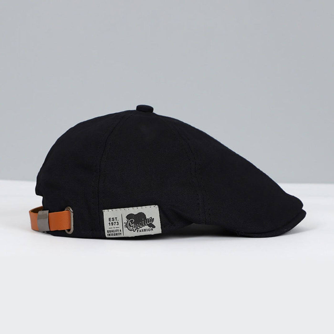 Black flat cap with sleek design, brown strap, and tag, set against a grey and white surface.