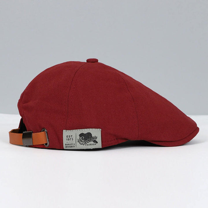 Red flat cap featuring a brown adjustable strap and stylish tag, on a muted grey background.