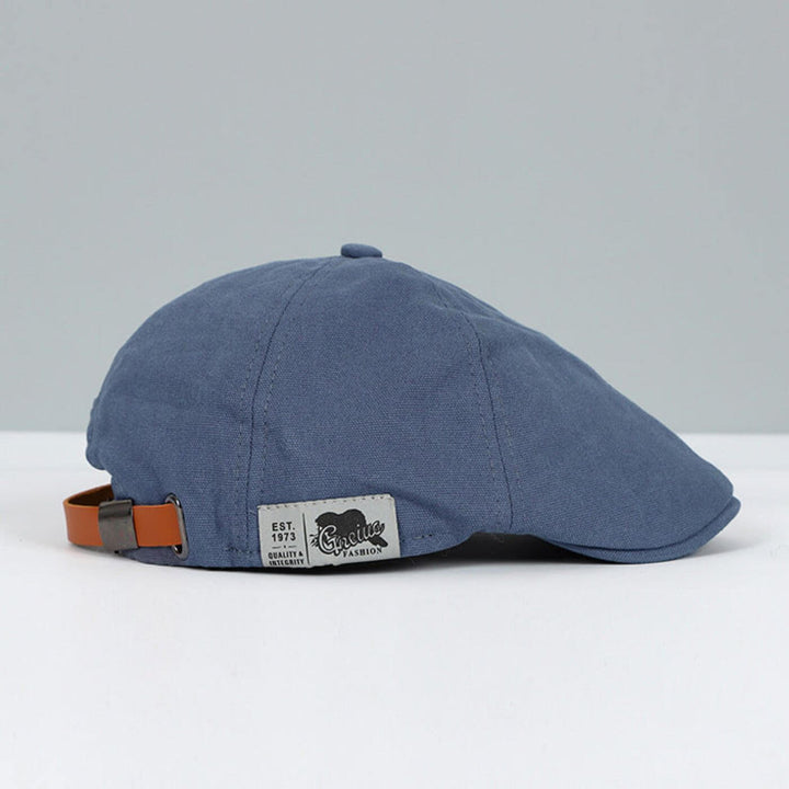 Blue flat cap with brown adjustable strap and stylish tag, on a grey and white surface.