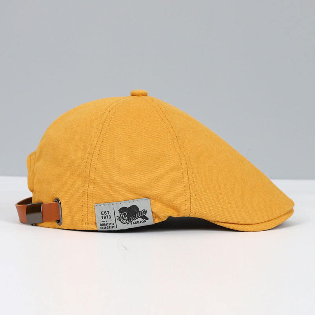 Mustard yellow flat cap featuring brown strap and tag, displayed on a light grey backdrop.