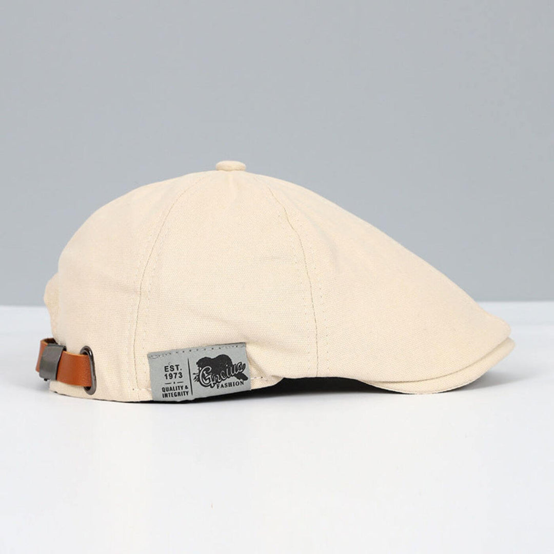 Beige flat cap with brown adjustable strap and stylish tag, placed on a light grey and white surface.