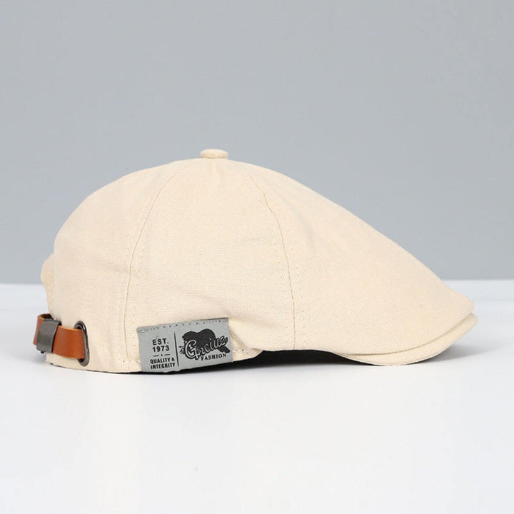 Beige flat cap with brown adjustable strap and stylish tag, placed on a light grey and white surface.