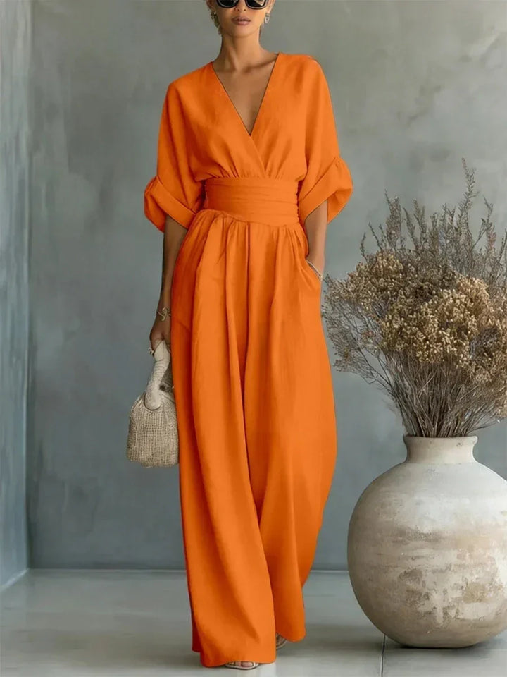 Orange V-neck high-waist jumpsuit featuring rolled-up sleeves, set in a studio with a minimalist vase and artistic foliage.