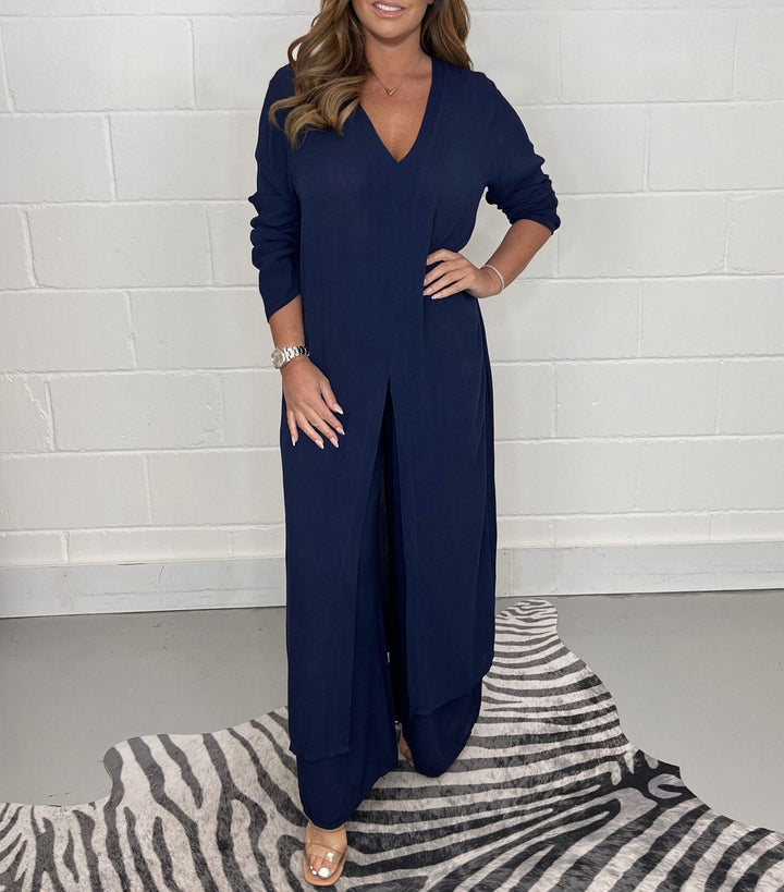 Model wearing a dark blue two-piece set with long sleeves and a V-neck, standing on a zebra-patterned rug against a white brick wall.