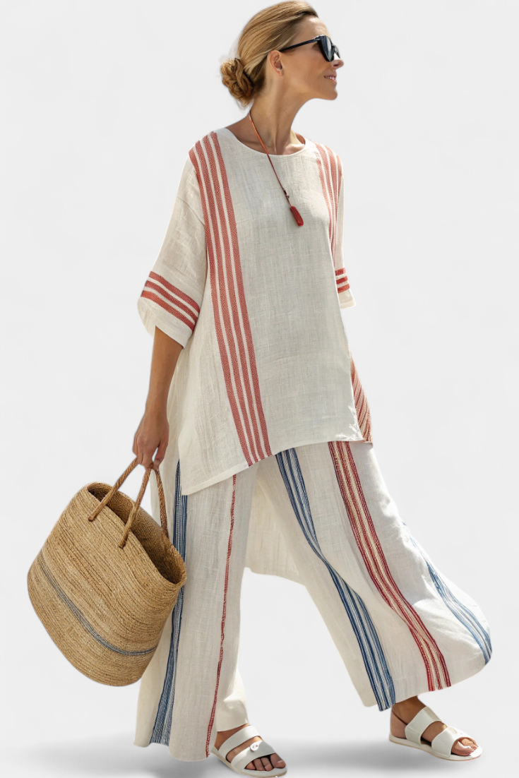 Woman in white two-piece set with red and blue stripes, holding large straw tote, sunglasses on, plain background.