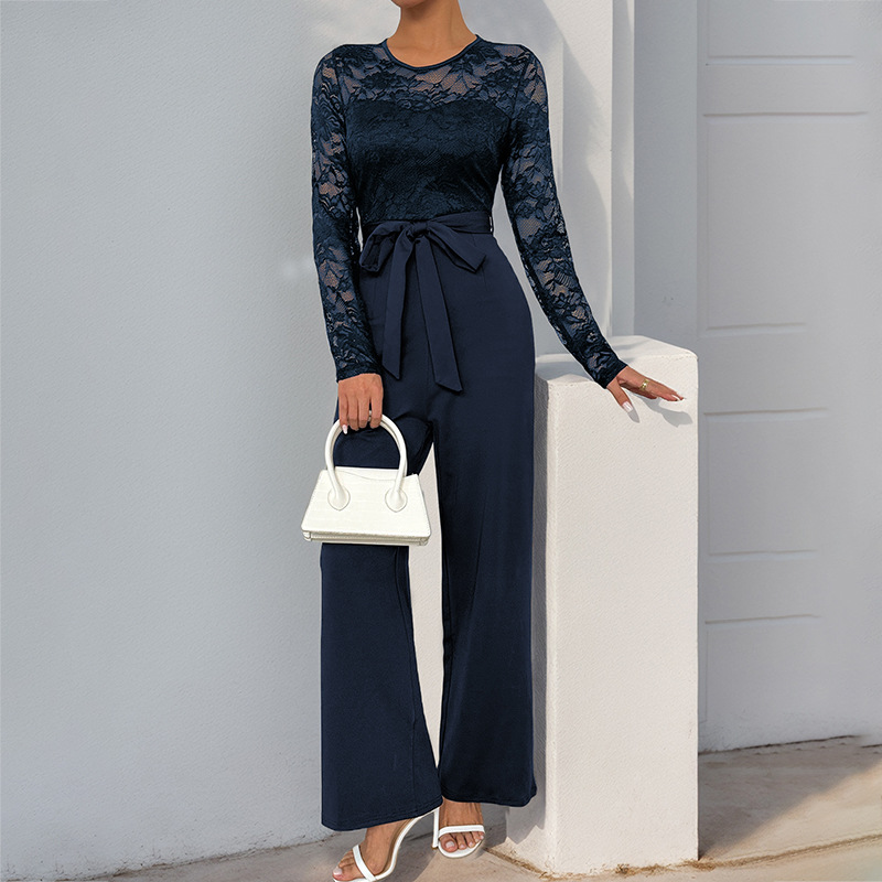 Navy lace bell sleeve jumpsuit with a waist tie, paired with a white handbag and sandals, outdoor light setting.