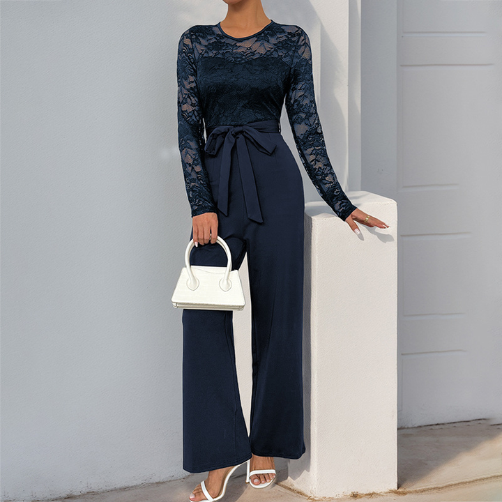 Navy lace bell sleeve jumpsuit with a waist tie, paired with a white handbag and sandals, outdoor light setting.