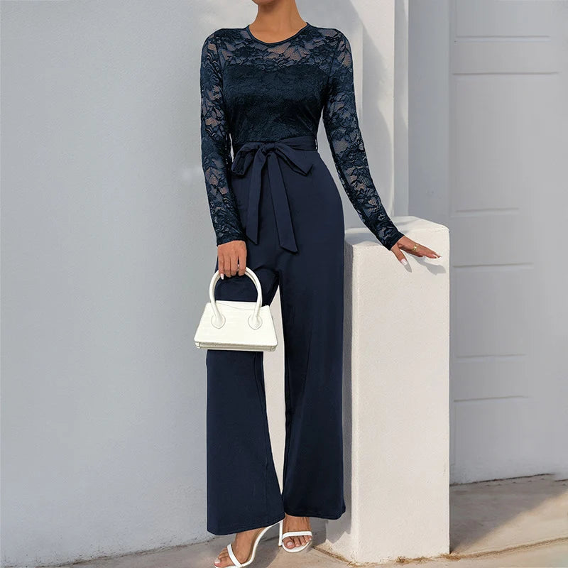 Elegant navy jumpsuit with lace sleeves, belted waist, white handbag, and sandals, posed outdoors by a light wall.