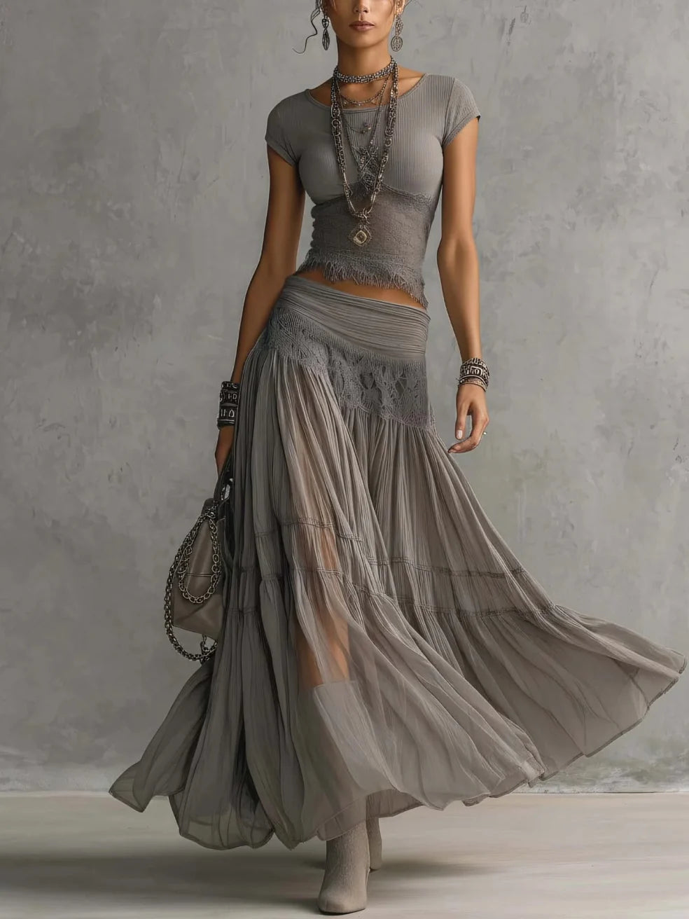 Woman in a gray maxi skirt set with lace details, wearing layered necklaces and bracelets, standing in a studio.