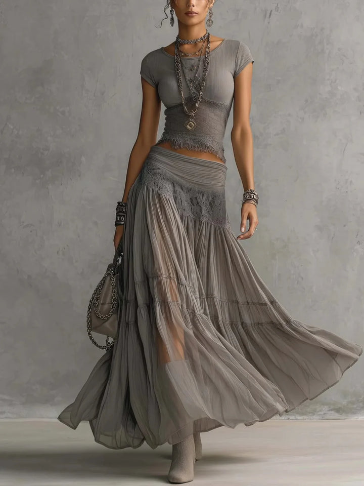Woman in a gray maxi skirt set with lace details, wearing layered necklaces and bracelets, standing in a studio.