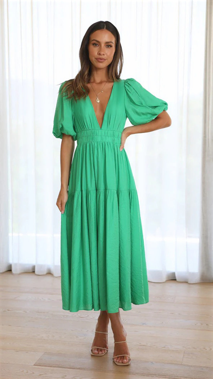 Woman wearing a green midi dress with puff sleeves, standing on wooden floor with sheer curtains.