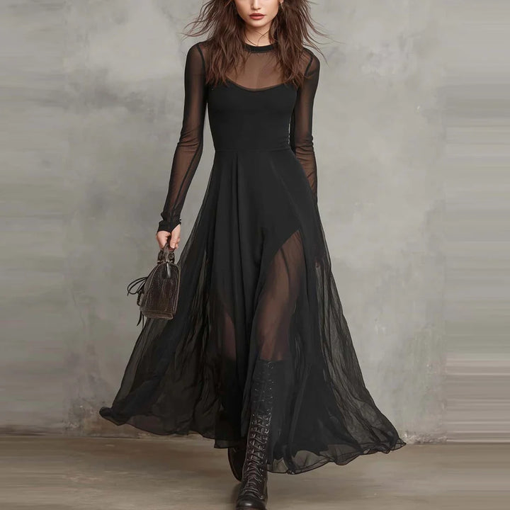 Woman wearing black maxi dress with sheer overlays, carrying small bag, standing in front of a gray textured wall.