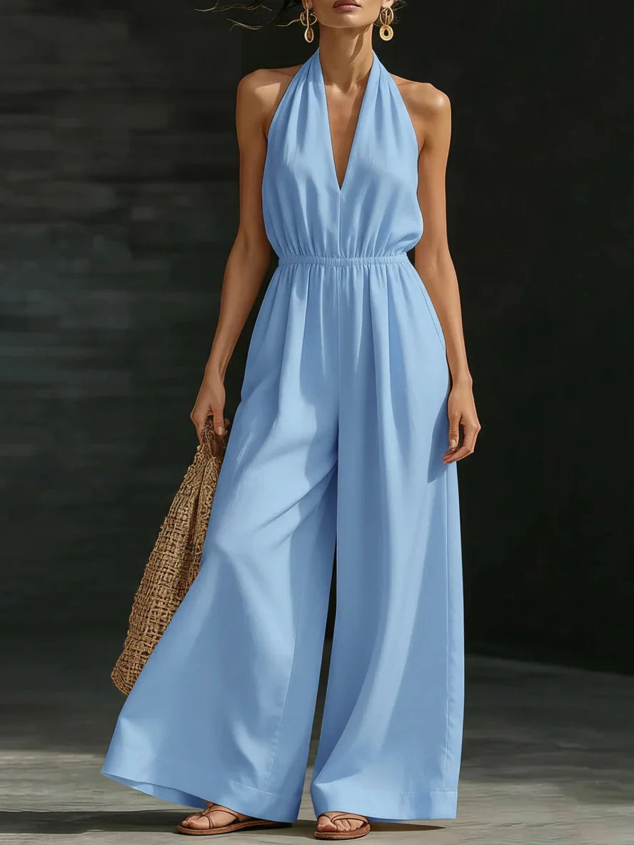 Woman in a light blue jumpsuit with halter neck and wide legs, carrying a woven bag, photographed in a dark studio.