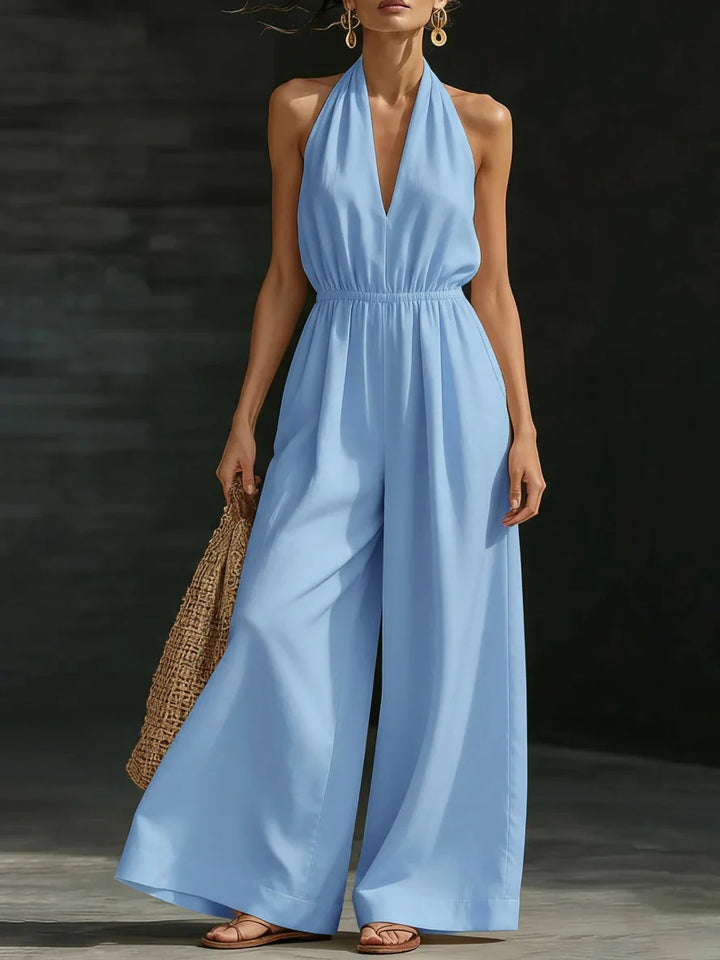 Woman in a light blue jumpsuit with halter neck and wide legs, carrying a woven bag, photographed in a dark studio.
