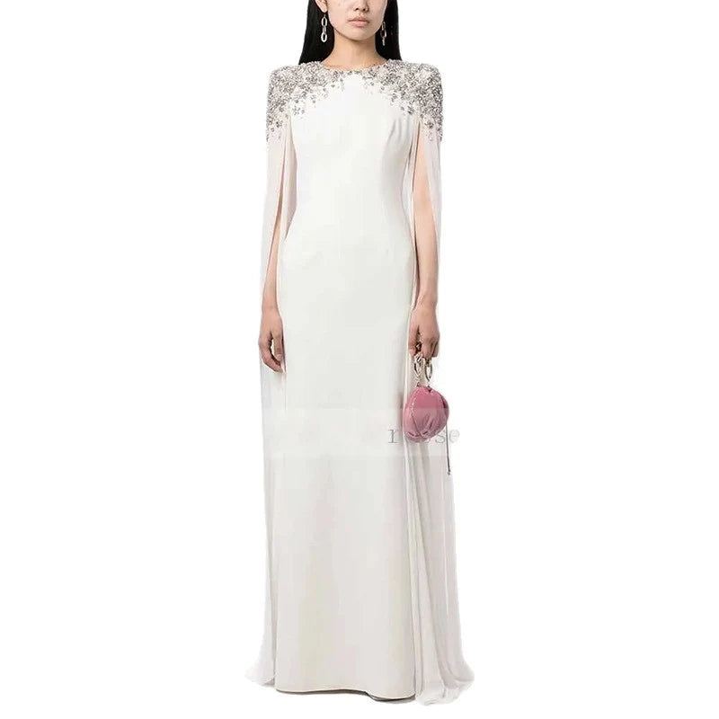 White elegant dress with sparkling shoulder details and sheer cape sleeves, holding a pink clutch in a studio.
