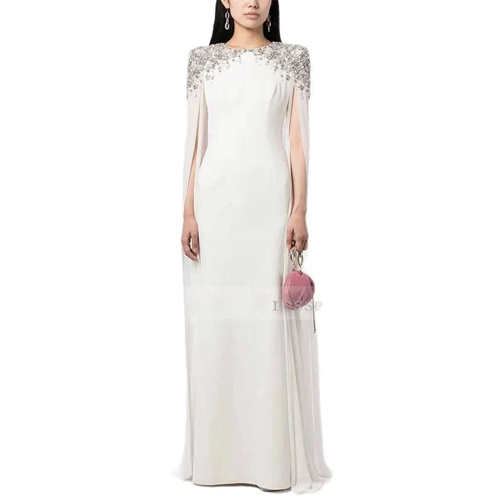White elegant dress with sparkling shoulder details and sheer cape sleeves, holding a pink clutch in a studio.