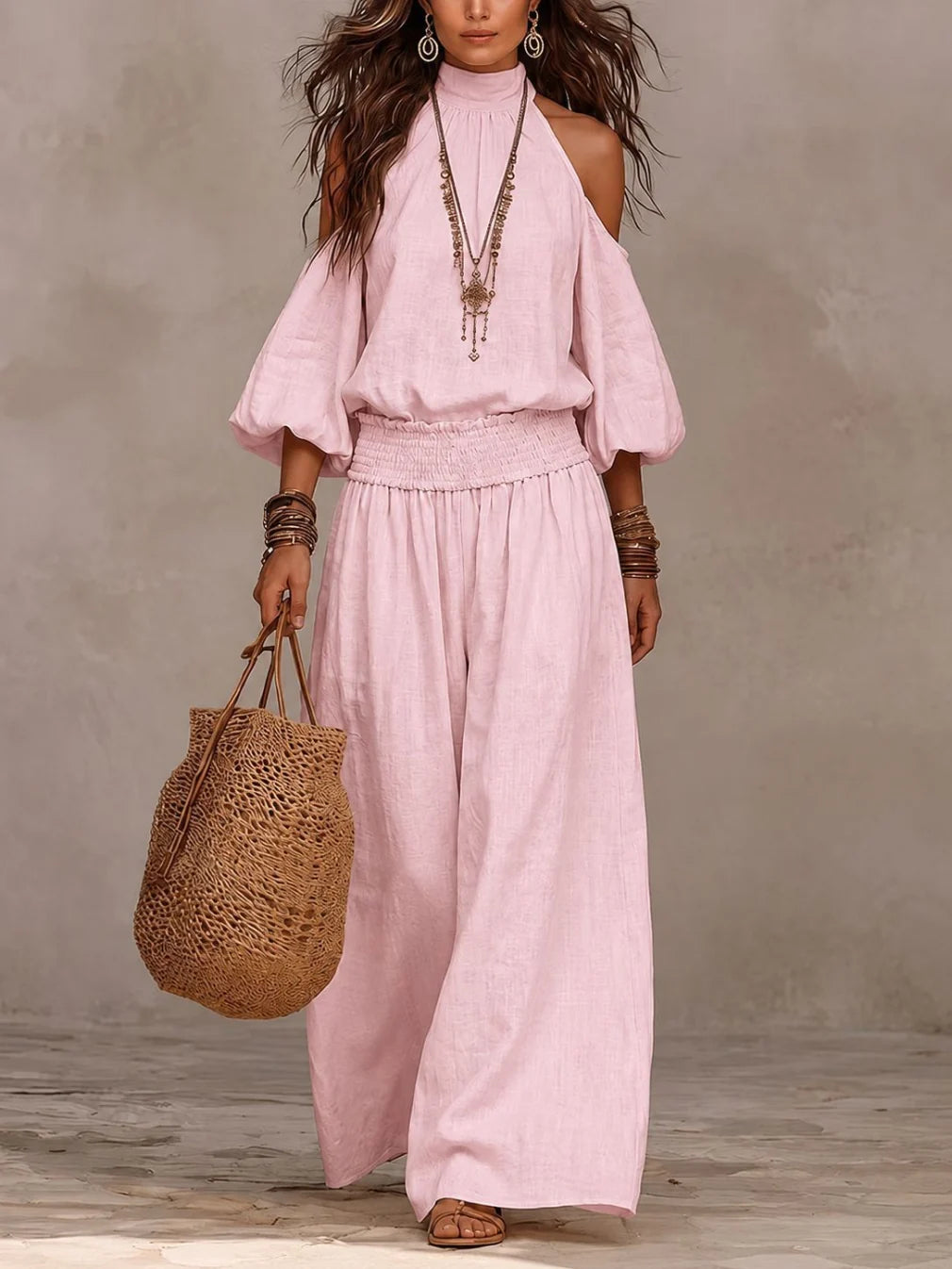 Model in pink off-shoulder jumpsuit, adorned with layered necklaces and holding a woven tote, against a plain background.