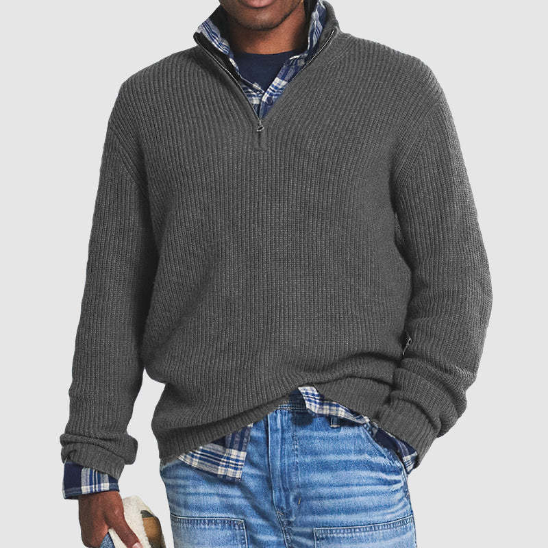 Man wearing a grey zip sweater over a plaid shirt with jeans, holding a hat in a studio setting.