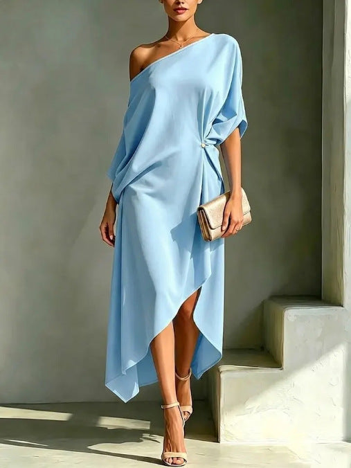 Elegant asymmetrical light blue robe with one-shoulder design, accessorized with a clutch and sandals in a minimal studio setting.