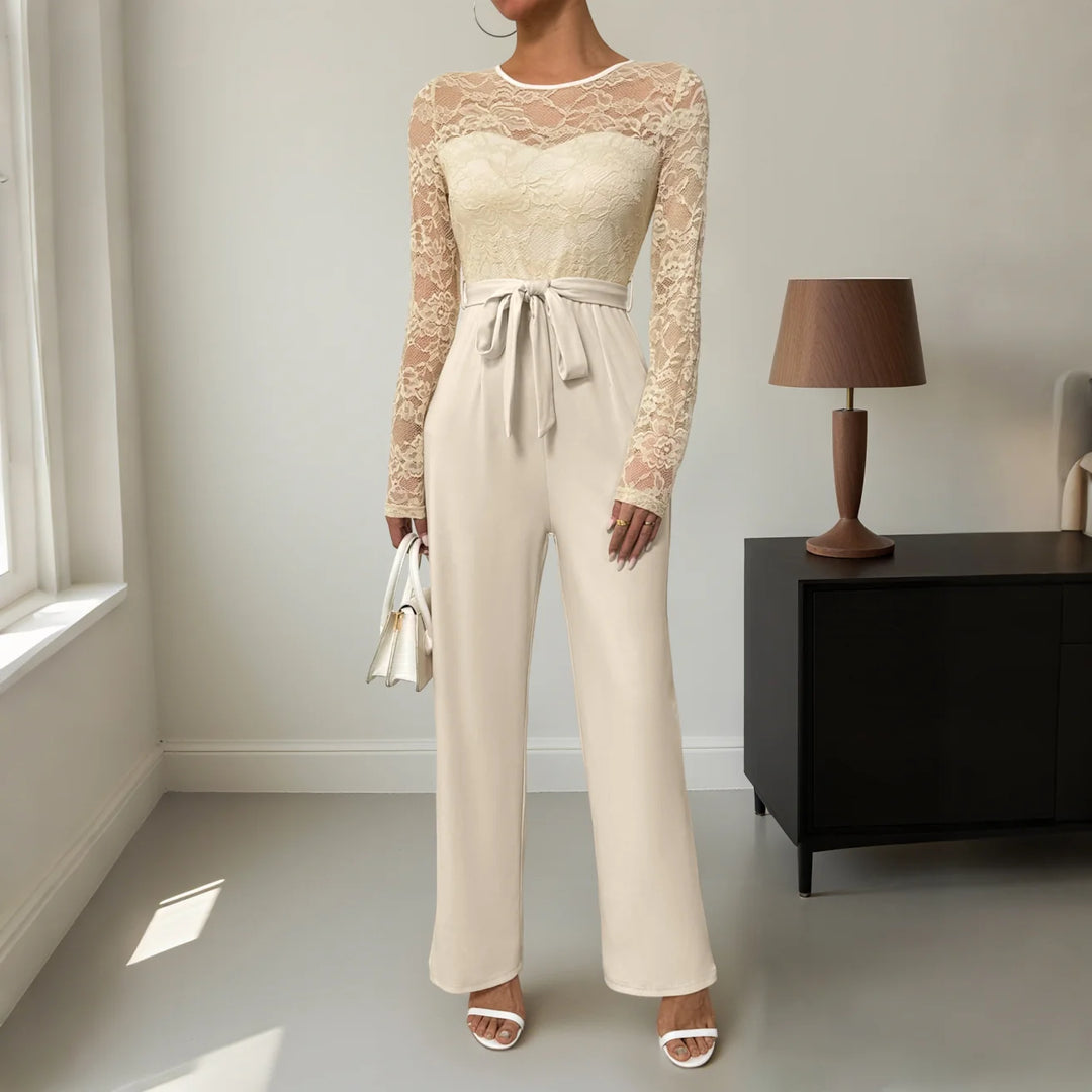Chic beige jumpsuit with lace long sleeves, tie waist, white handbag, indoor room.
